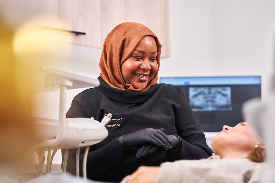 A photograph of a dental clinician smiling at a patient laying in a dental chair with an image of an x-ray on a computer screen behind them