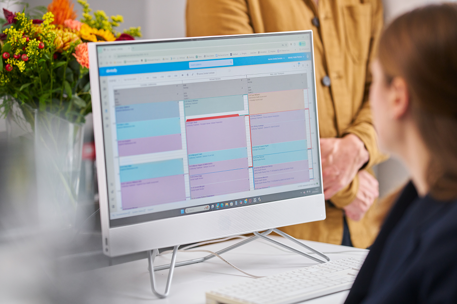 A photograph of a receptionist looking at a computer with the Dentally software on screen displaying a calendar